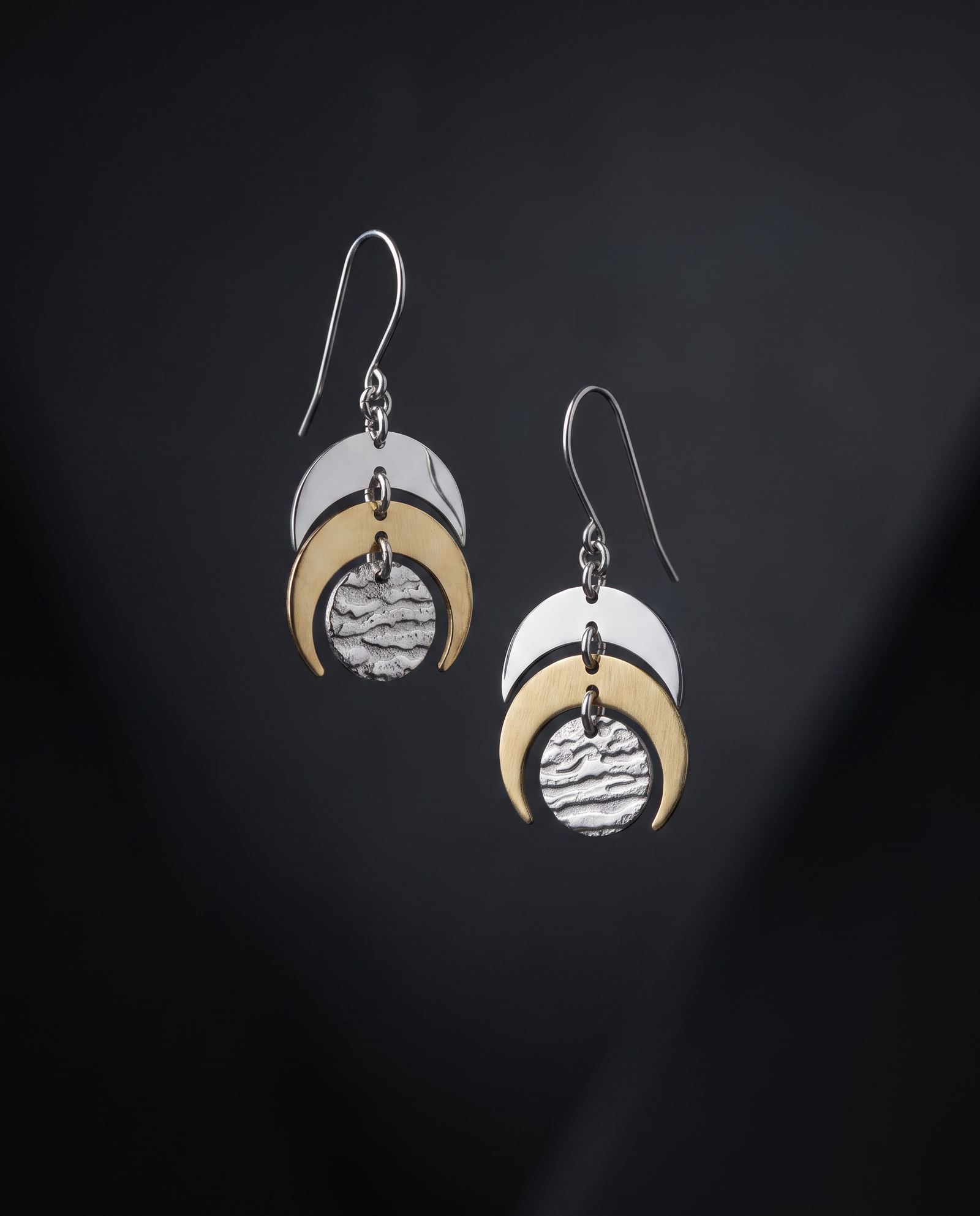 A pair of silver and gold crescent moon dangle earrings with textured circular charms, displayed against a dark black background.