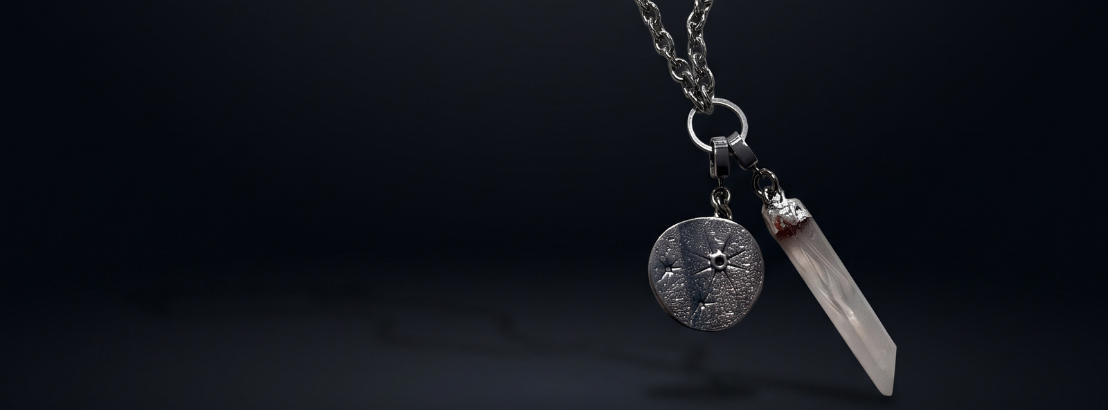 A silver-toned chain necklace featuring a round textured medallion and a pointed clear crystal pendant, displayed against a dark gradient background.
