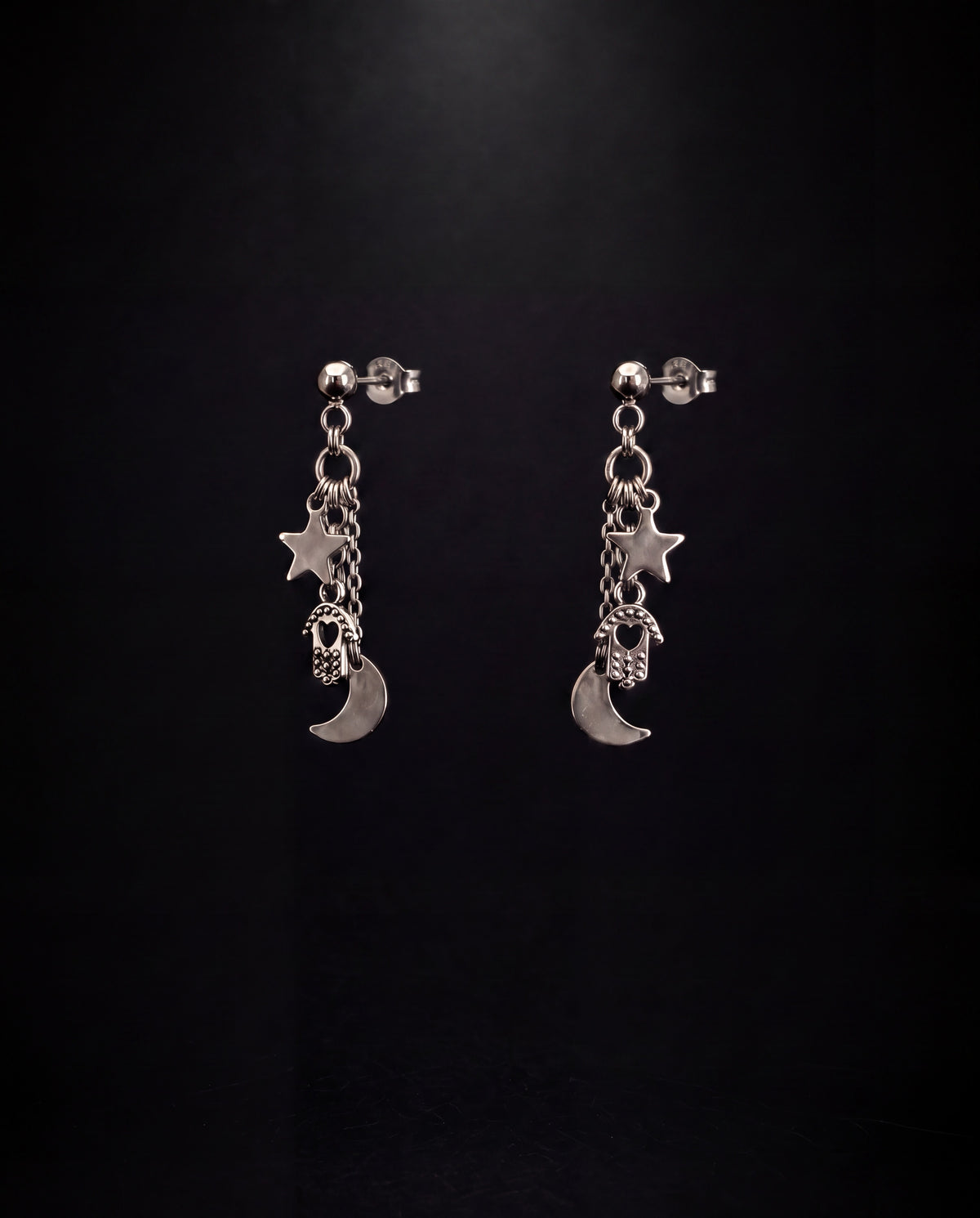 A pair of silver-toned dangle earrings with star, crescent moon, and skull-and-bone charms, displayed against a dark black background.