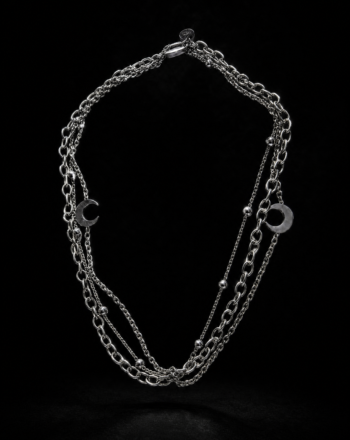 A multi-strand silver chain necklace with crescent moon and oval charms, displayed against a solid black background.