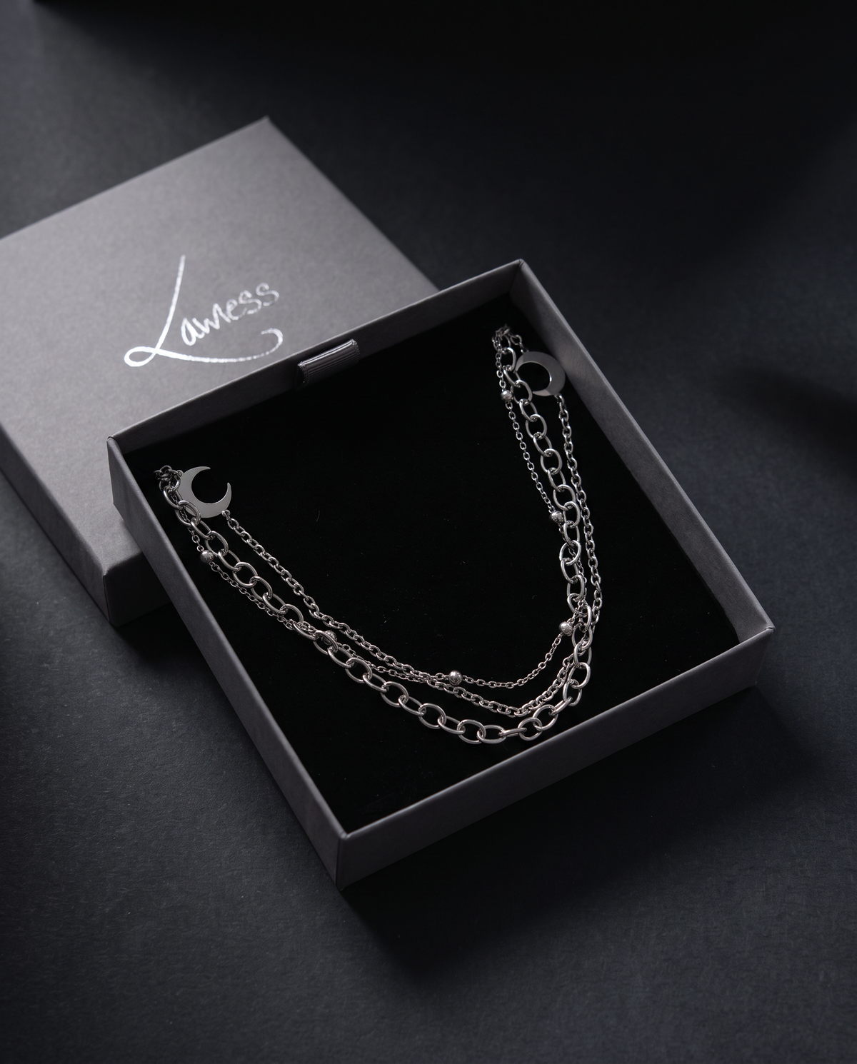 A silver-toned double-layer chain bracelet with a crescent moon charm, presented in an open dark gray jewelry box with a black velvet lining from lawless Jewellery.