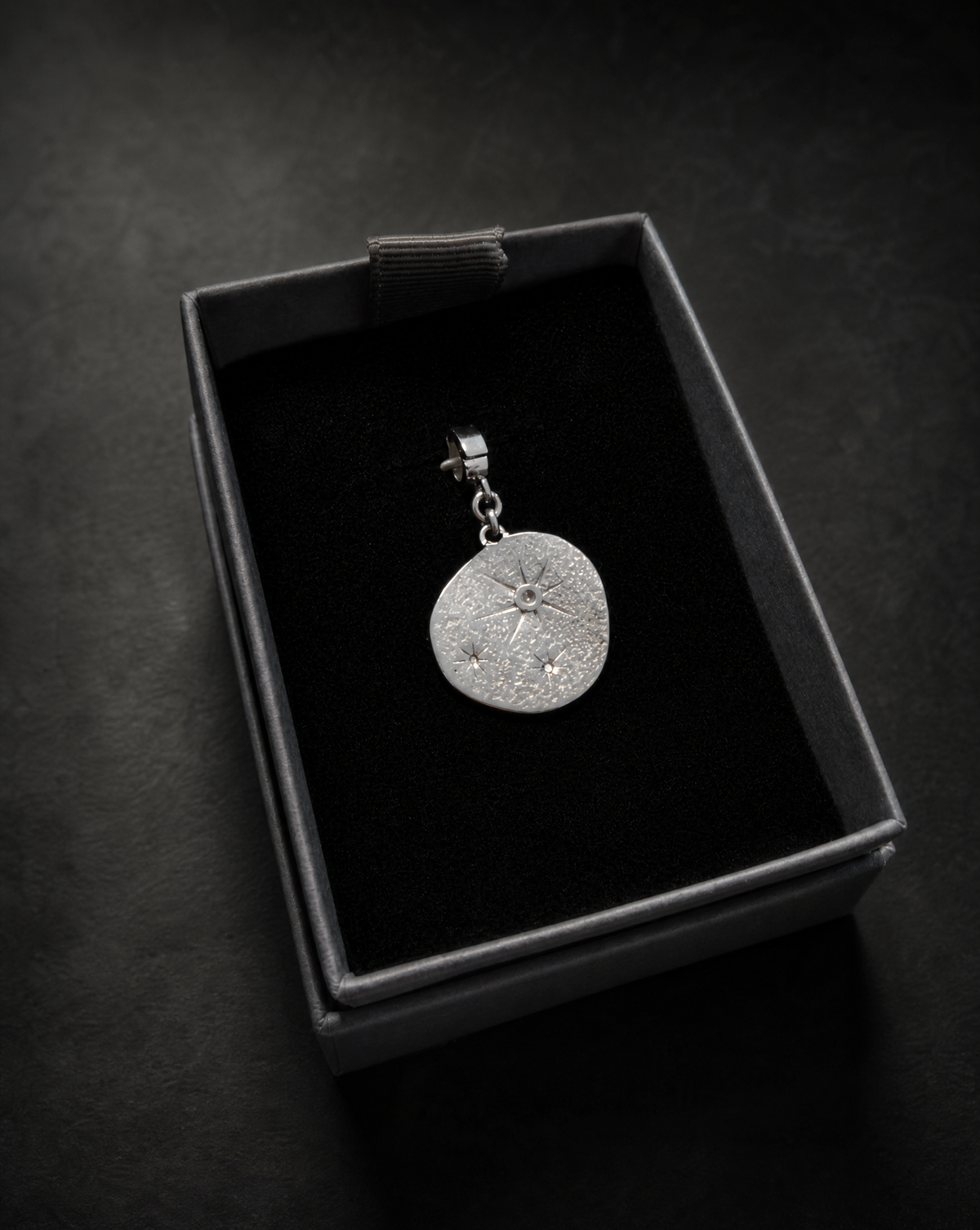 Silver Hecate Star Gazer Charm displayed in a black jewellery box, featuring a round textured disc with a radiating central star and two smaller celestial details.
