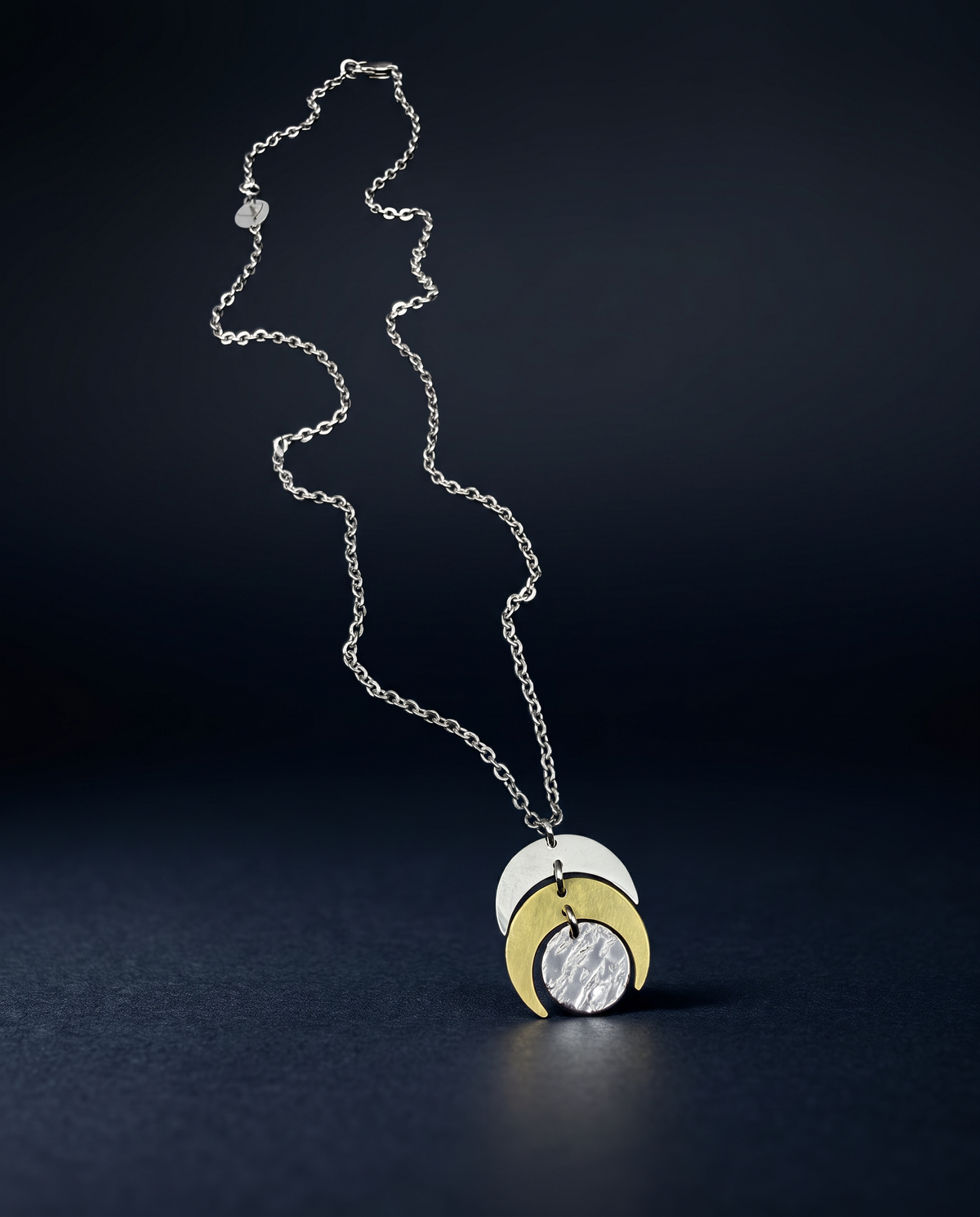 A silver chain necklace with a three-moon-phase pendant in polished silver and yellow gold, displayed on a dark, reflective surface.