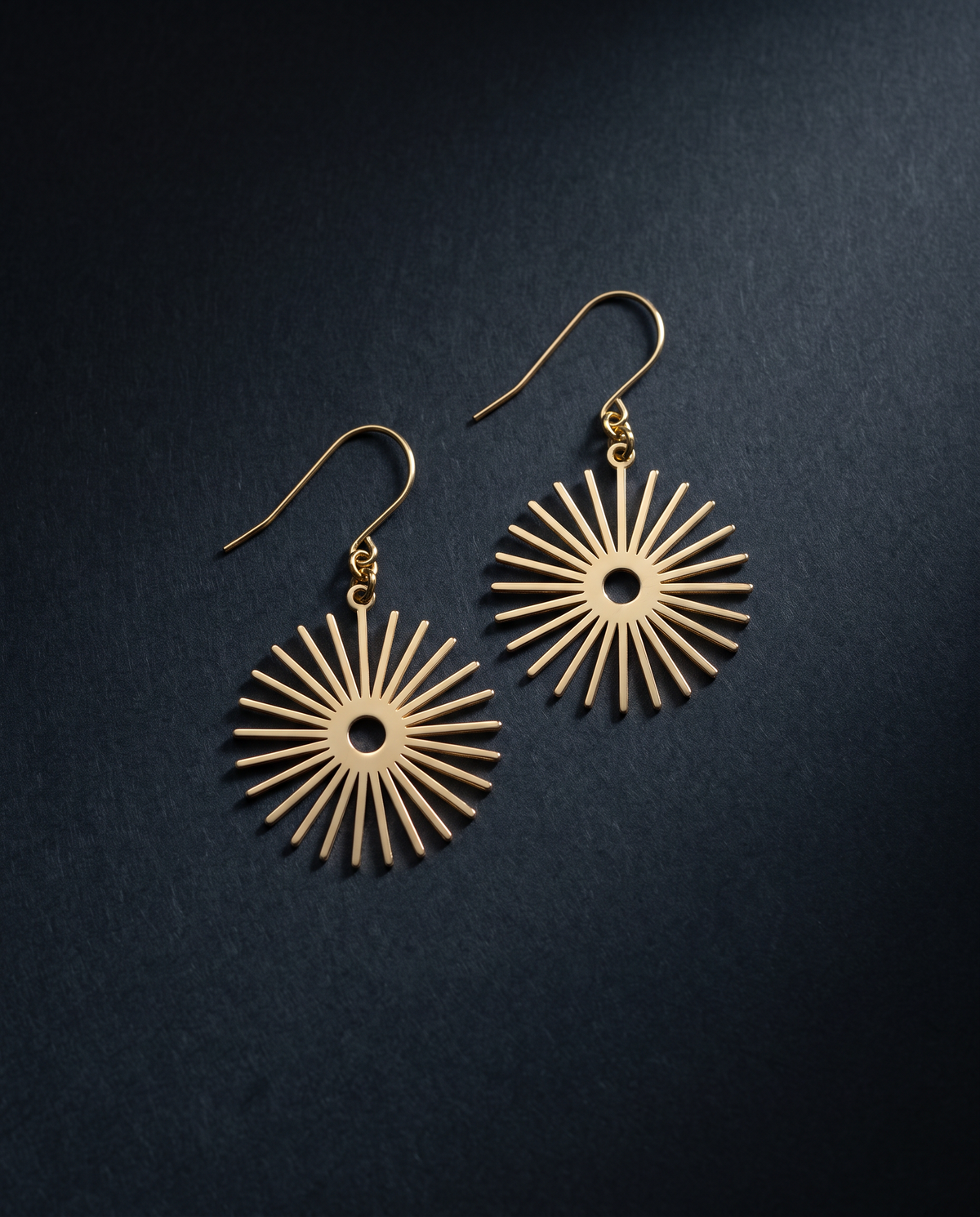 A pair of gold-toned dangle earrings with a sunburst or starburst radial disc design, displayed on a dark textured background.