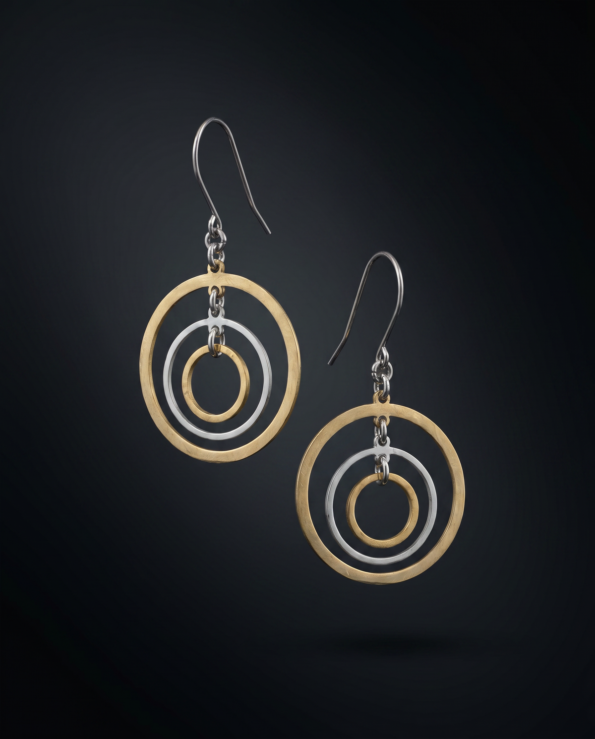 A pair of geometric dangle earrings featuring concentric gold and silver-tone circular rings suspended from silver fishhook wires, set against a dark gradient background.