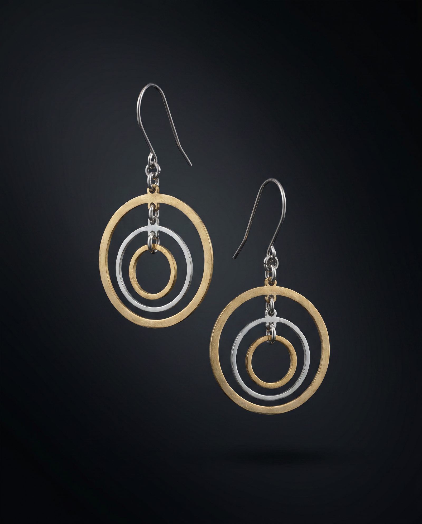 A pair of geometric dangle earrings featuring concentric gold and silver-tone circular rings suspended from silver fishhook wires, set against a dark gradient background.