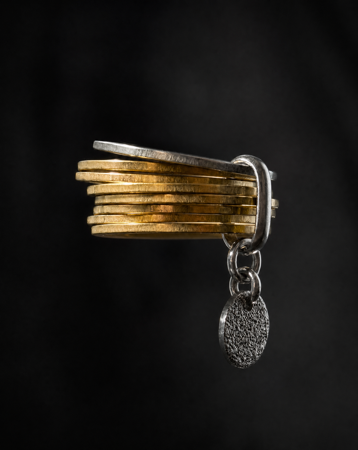 A stack of gold coins bound together by a silver clasp, with a textured silver oval pendant hanging from the clasp, set against a dark black background.