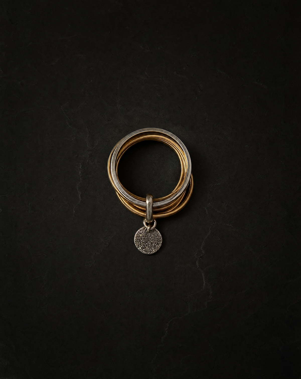 A stack of three multi-band rings in silver and gold tones with a small textured silver disc charm, displayed on a dark textured background.