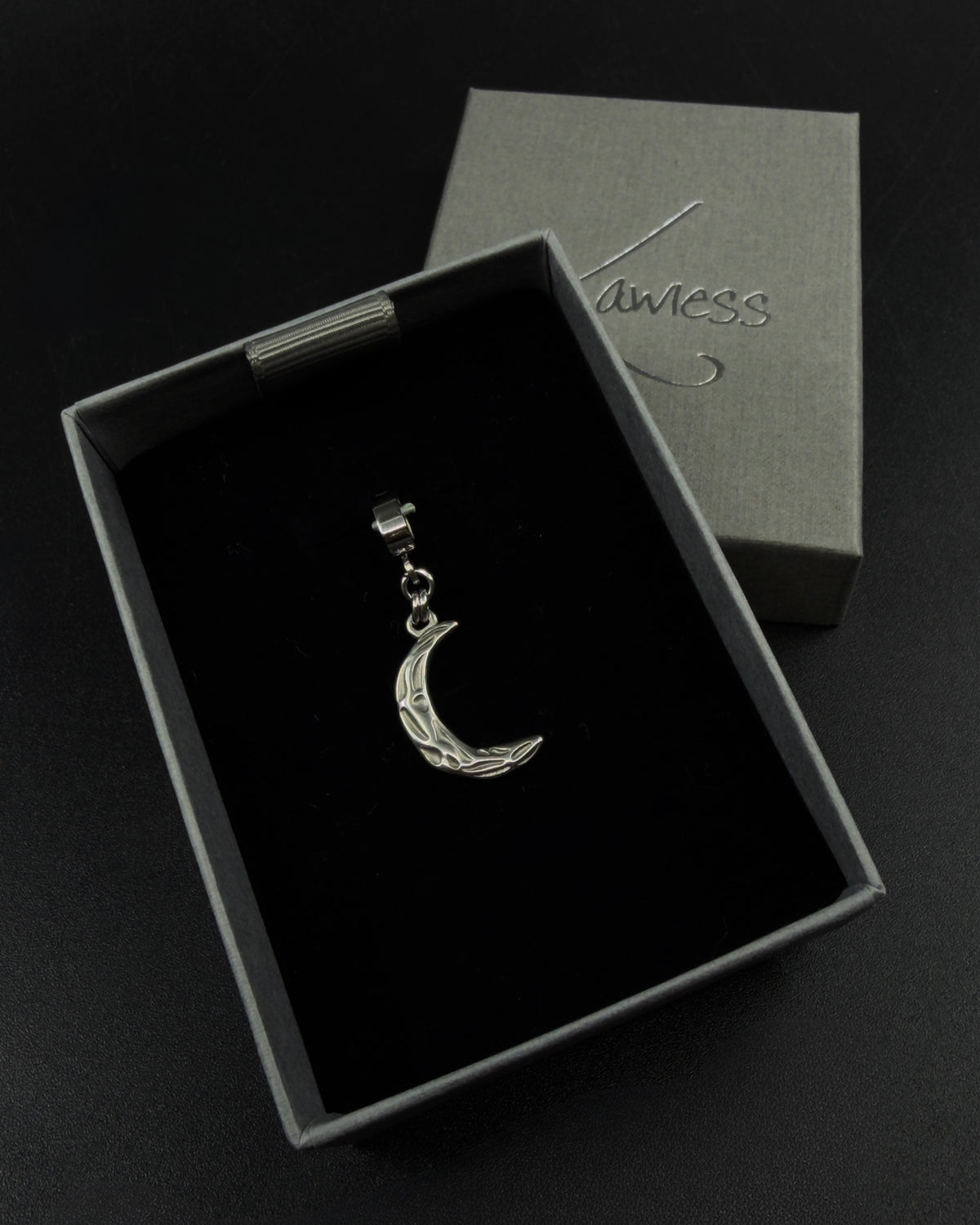 Silver Hecate Keeper Charm with a sculptural crescent shape, raised organic markings and a wide stainless steel bail against a black background.