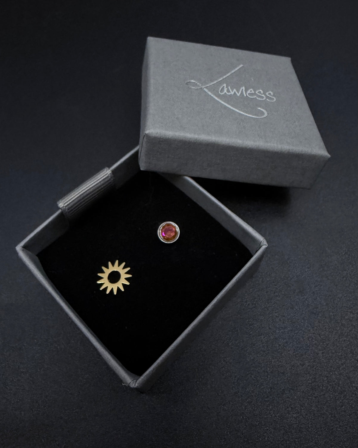 Two earrings, one gold and one silver, in a 'Lawless' branded box on a dark background.