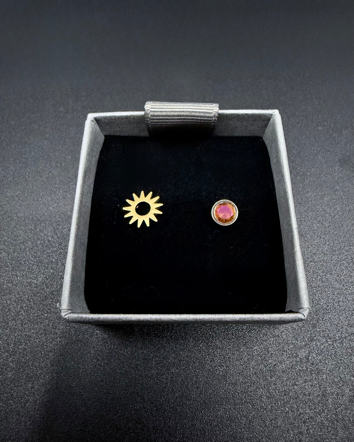 Two small earrings, one gold and one pink, in a jewelry box on a dark background.