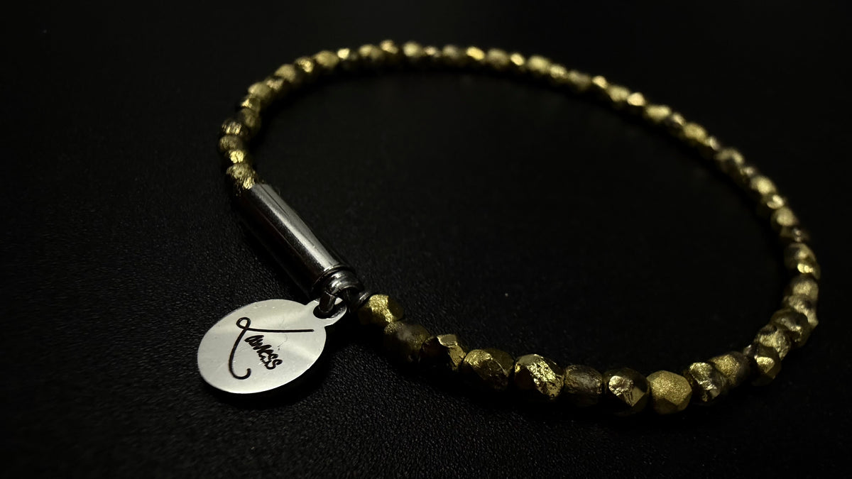 Gold Eos Flare Bracelet made from faceted burnished-gold crystal beads with a silver screw clasp and round Lawless logo tag against black fabric.
