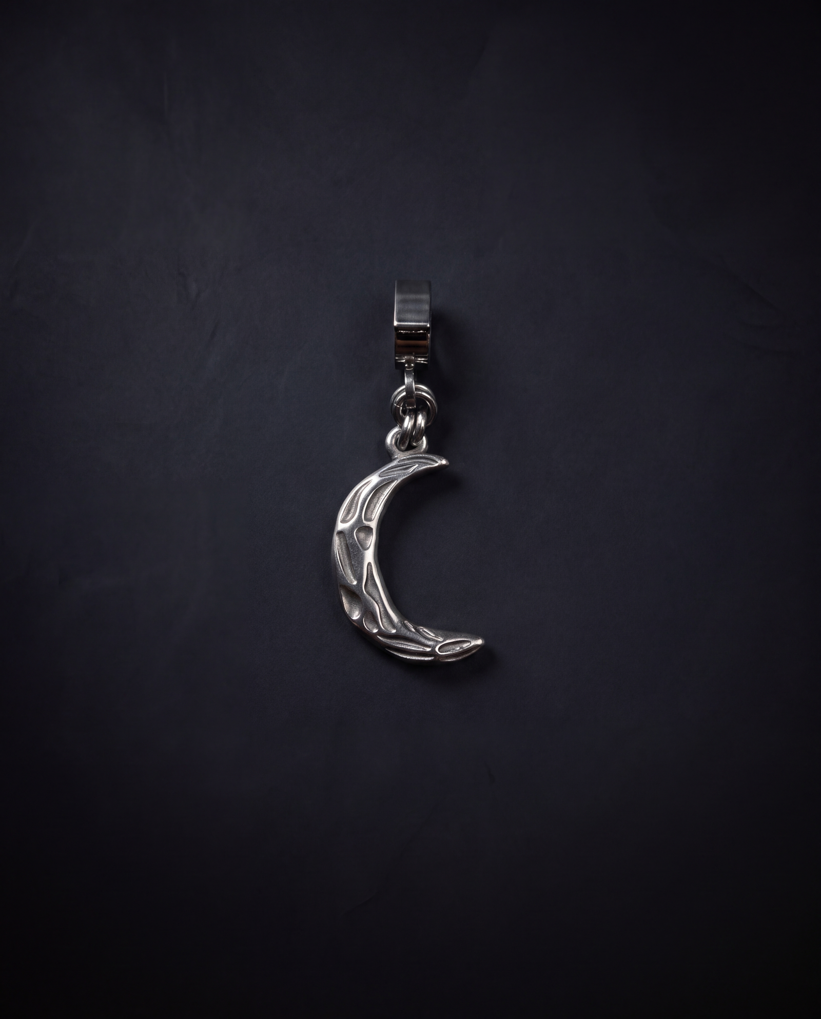 A silver crescent moon pendant charm with a textured, hammered finish, hanging from a matching silver ring on a dark, textured background.
