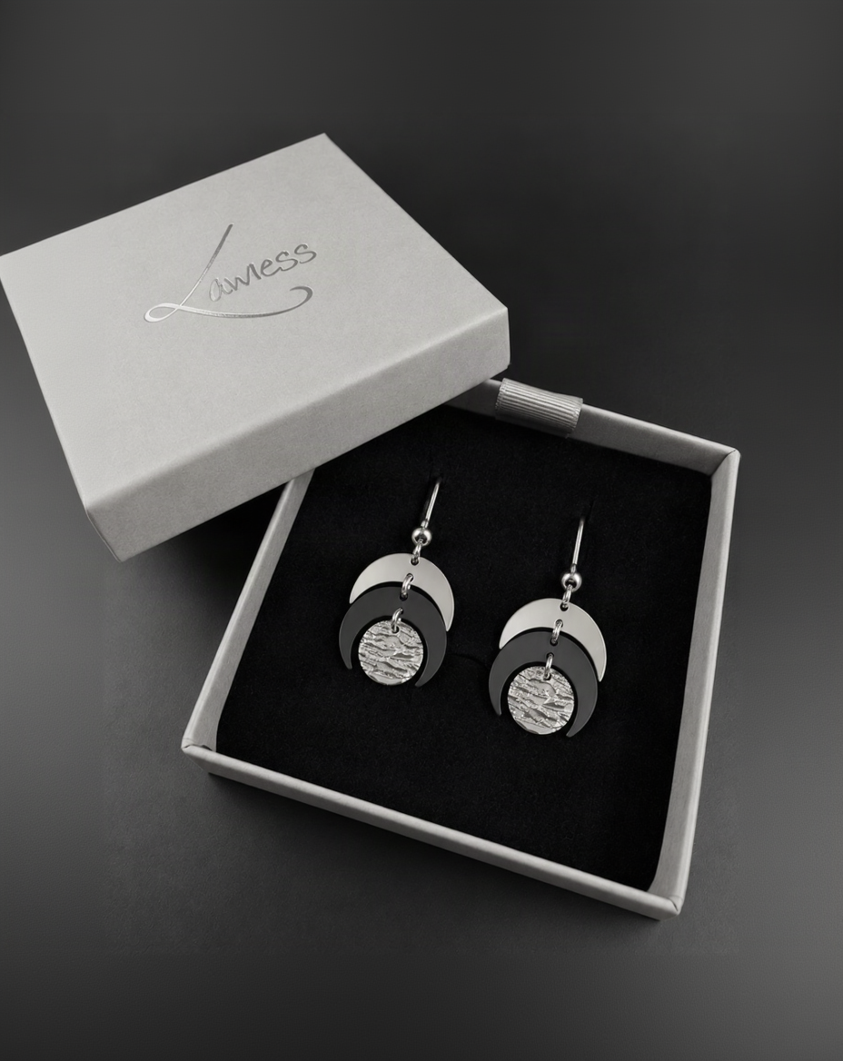 Pair of Luna Phase drop earrings with polished silver crescent forms and a textured black central disc suspended from silver hook ear wires.