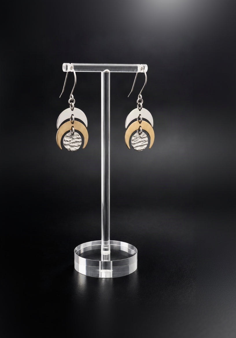 Luna Phase drop earrings with silver hooks, layered silver and gold crescents and textured silver lunar discs against a dark background.