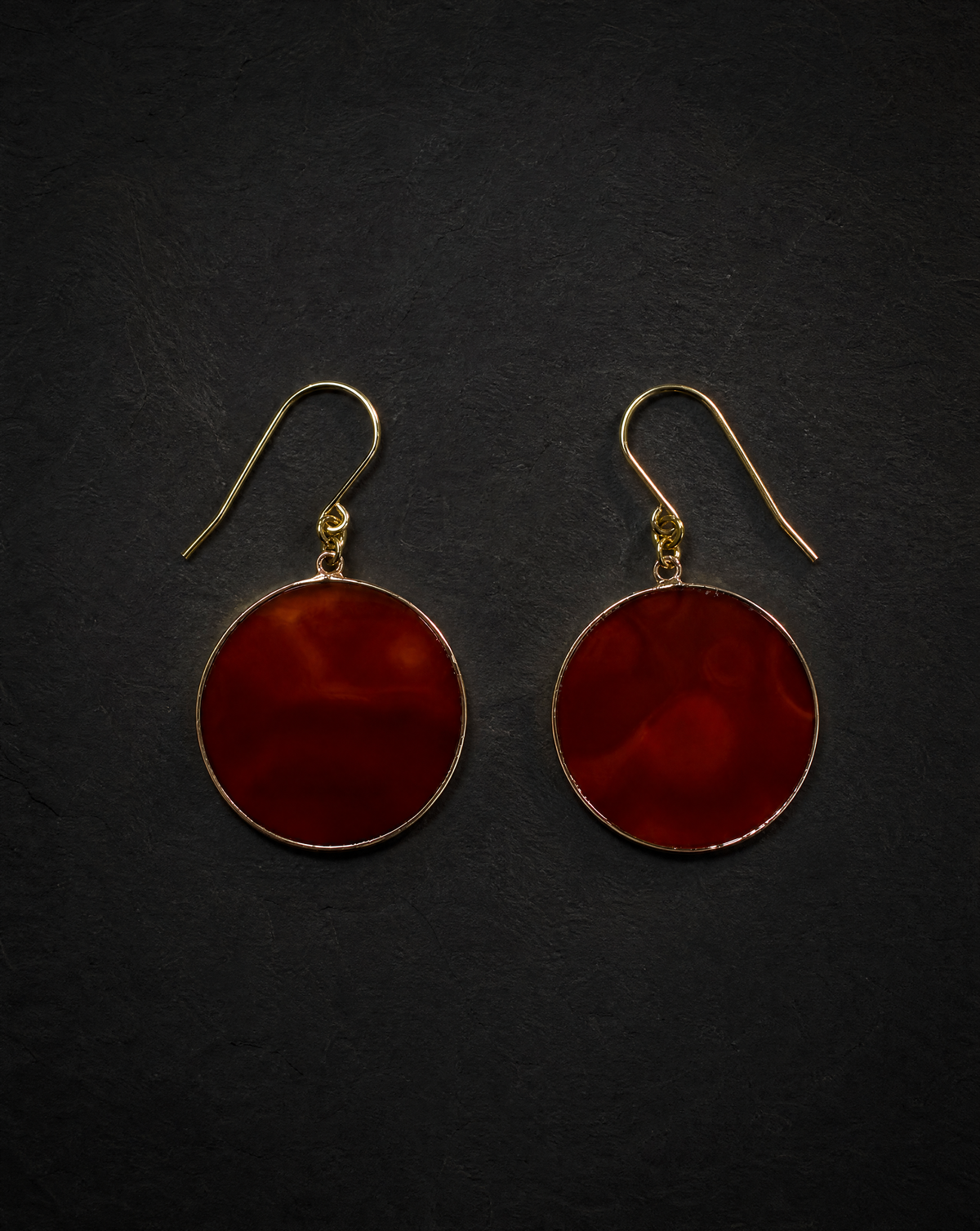 A pair of round drop earrings with deep red marbled stone centers and gold-tone hooks, displayed on a dark textured background.