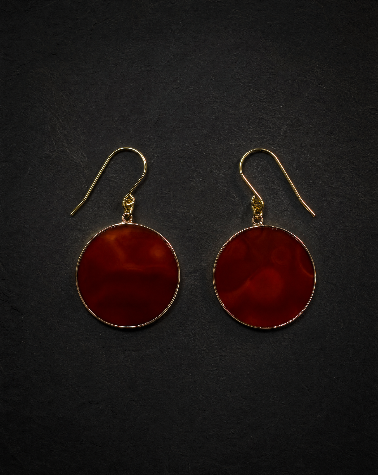 A pair of round drop earrings with deep red marbled stone centers and gold-tone hooks, displayed on a dark textured background.