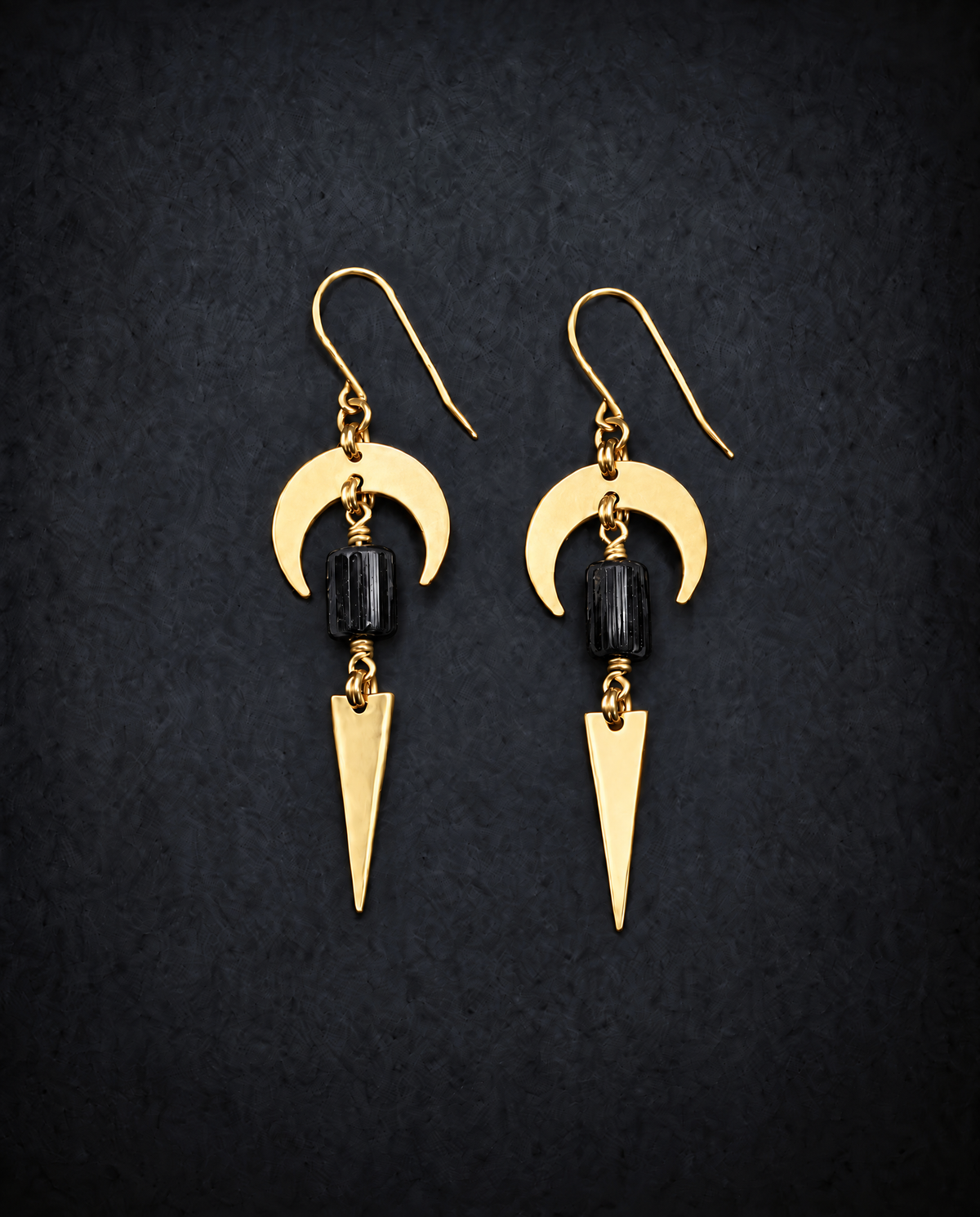 Pair of gold Orion Arc drop earrings with crescent shapes, rectangular black tourmaline gemstones and long triangle pendants against a dark background.