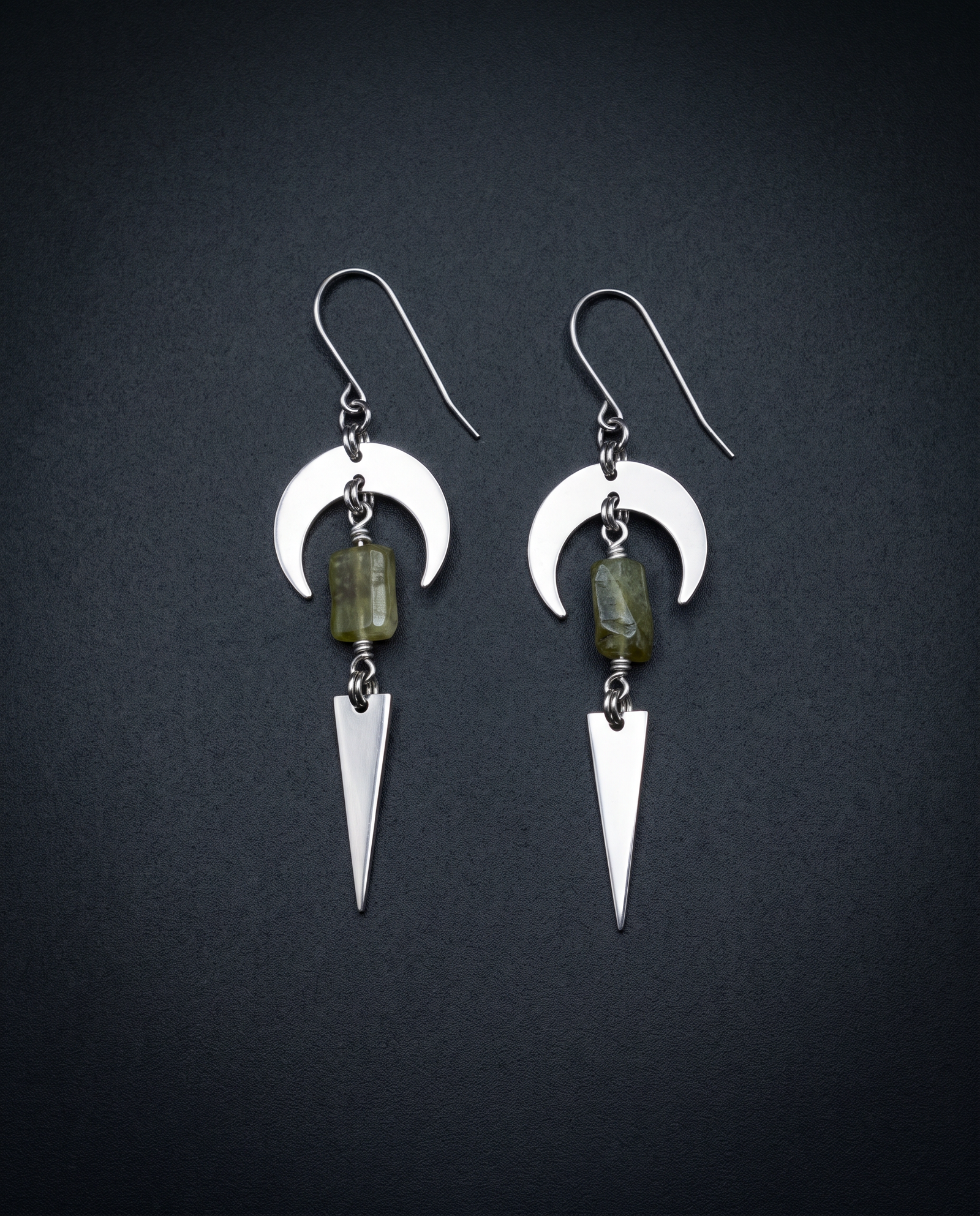 Pair of silver Orion Arc drop earrings with crescent shapes, natural green garnet stones and long triangle pendants against a dark background.