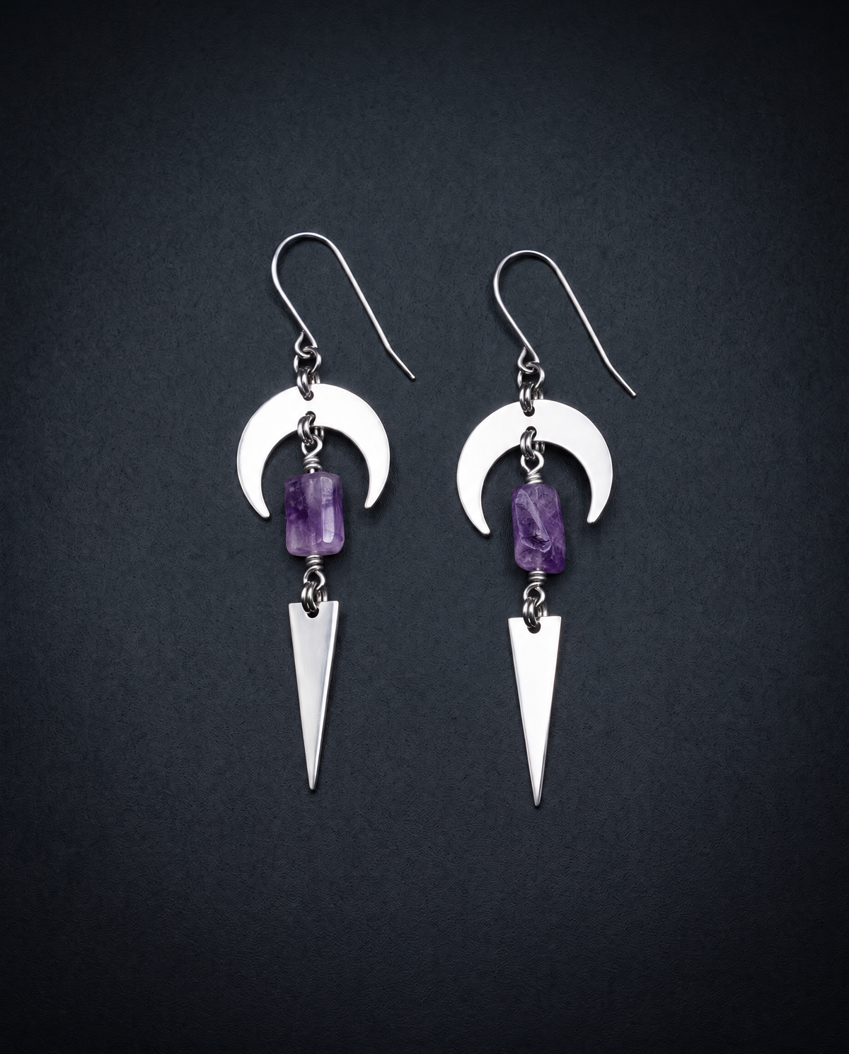 Pair of silver Orion Arc drop earrings with crescent shapes, rectangular violet amethyst stones and long triangle pendants against a dark background.