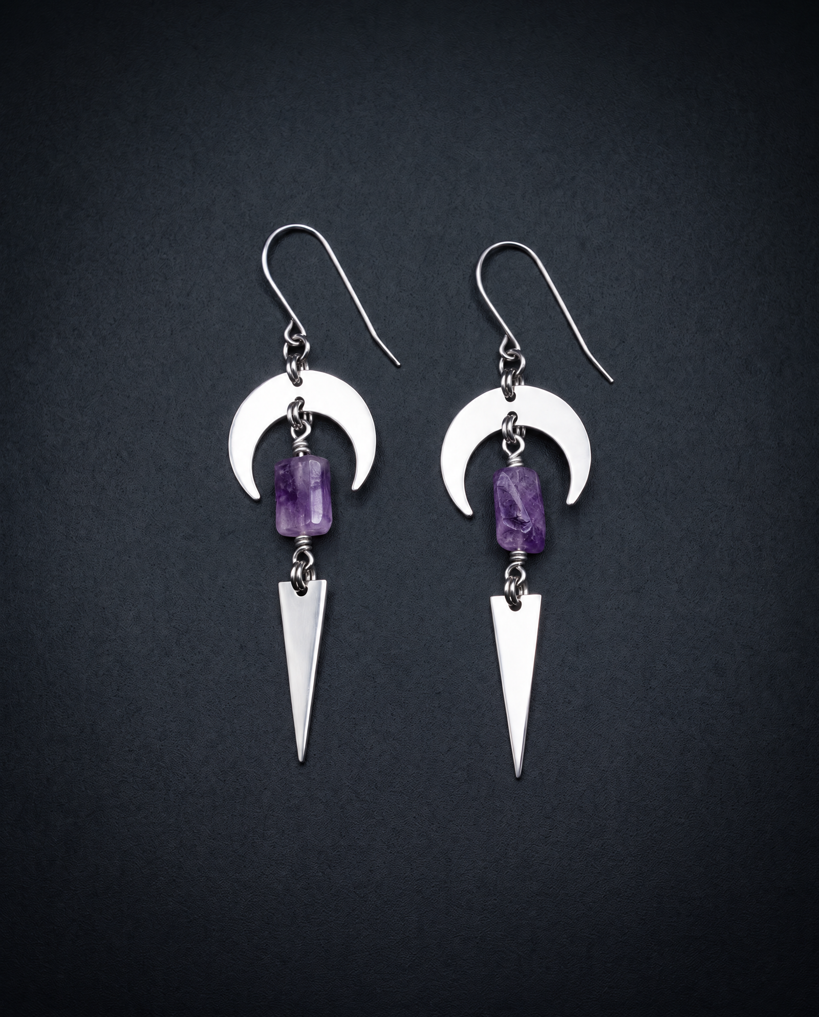 Pair of silver Orion Arc drop earrings with crescent shapes, rectangular violet amethyst stones and long triangle pendants against a dark background.