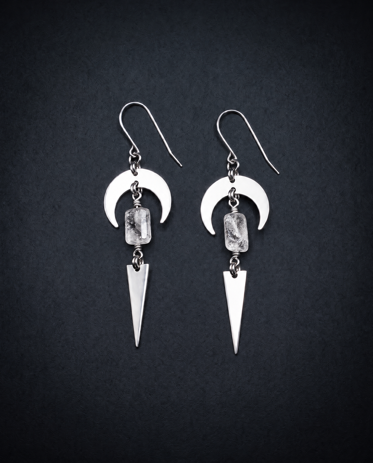 Pair of silver Orion Arc drop earrings with crescent shapes, natural clear quartz crystal stones and long triangle pendants against a dark background.