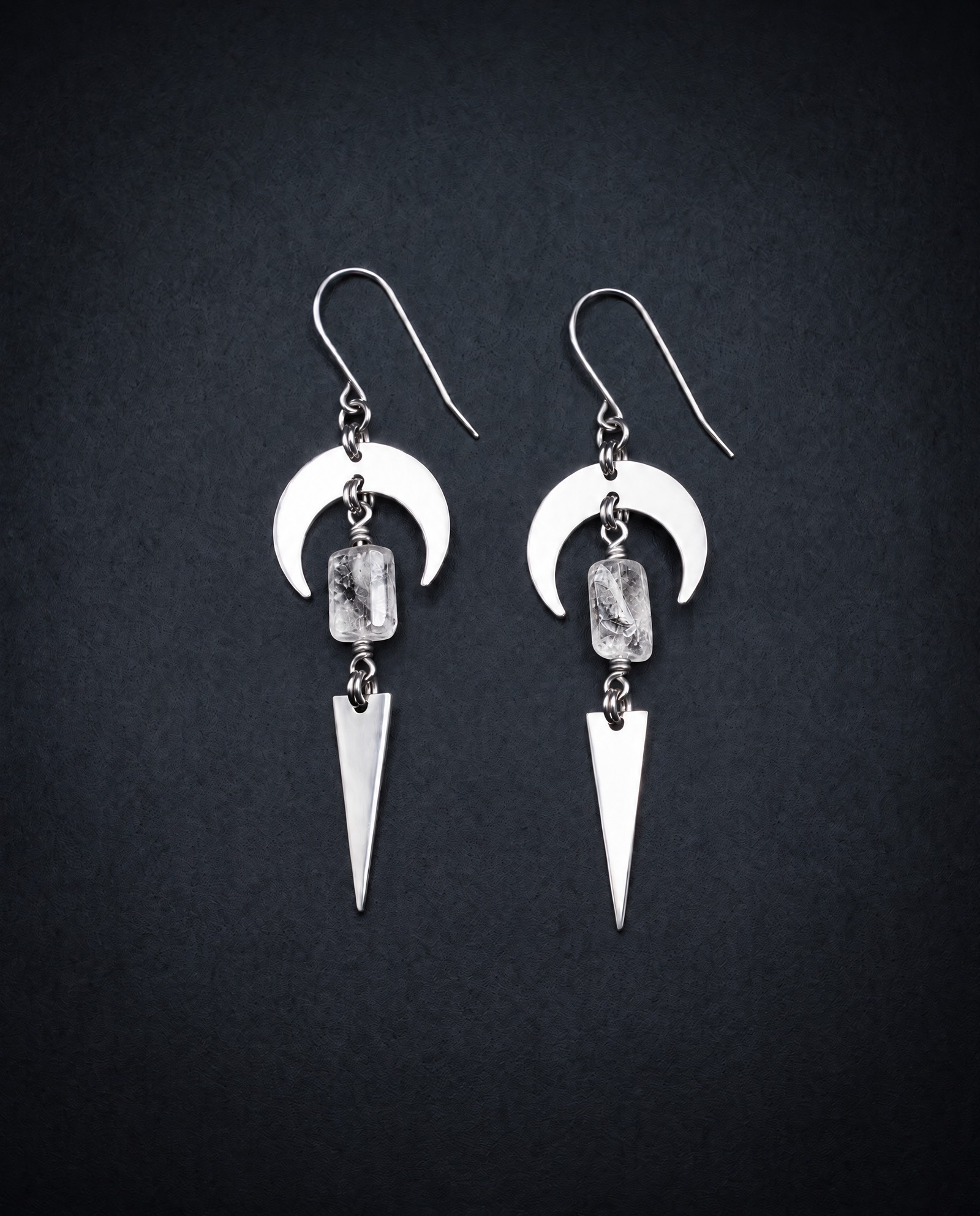 Pair of silver Orion Arc drop earrings with crescent shapes, natural clear quartz crystal stones and long triangle pendants against a dark background.