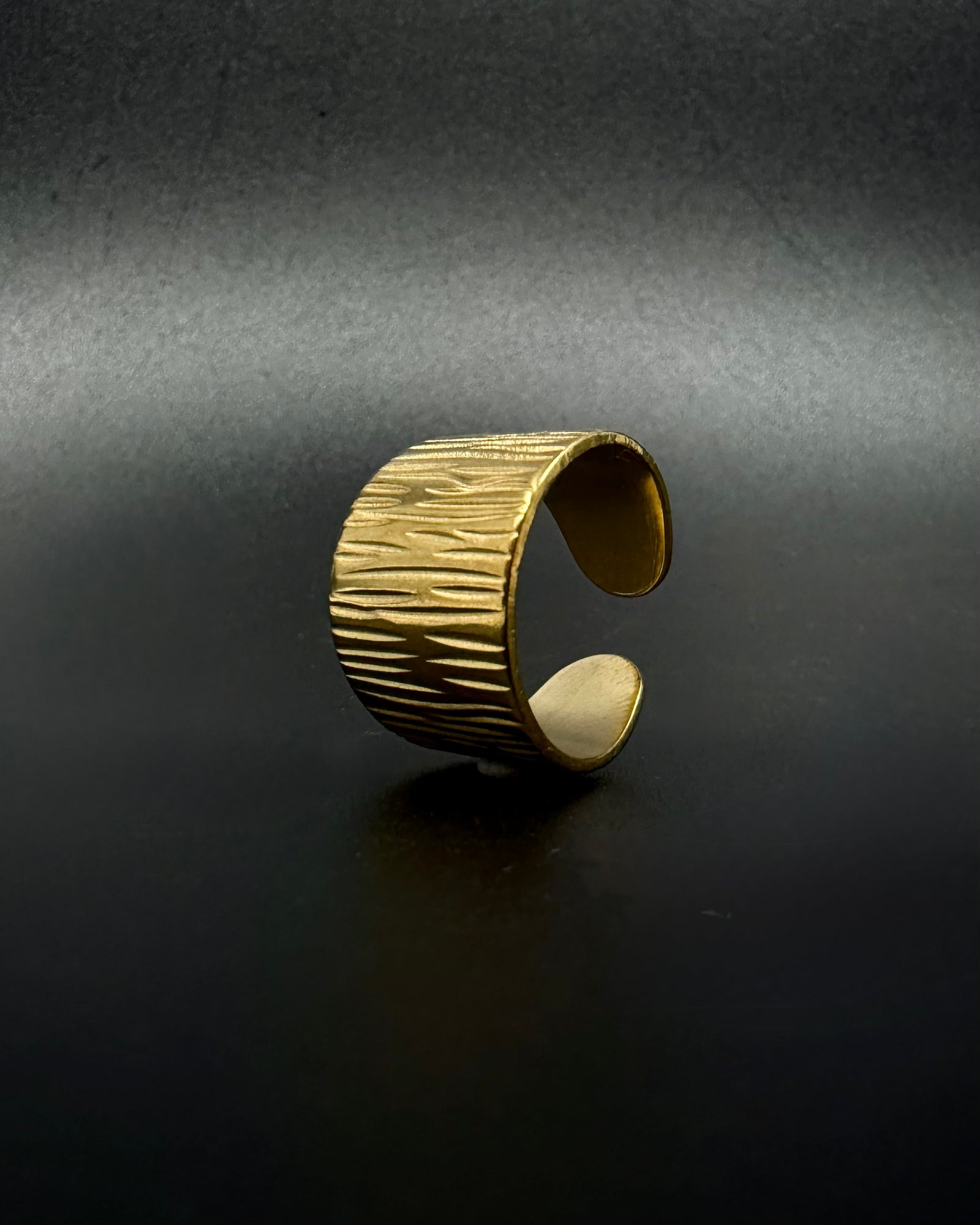 Gold Theia Frequency adjustable ring with a wide tapered band and irregular linear waveform texture against a black background.