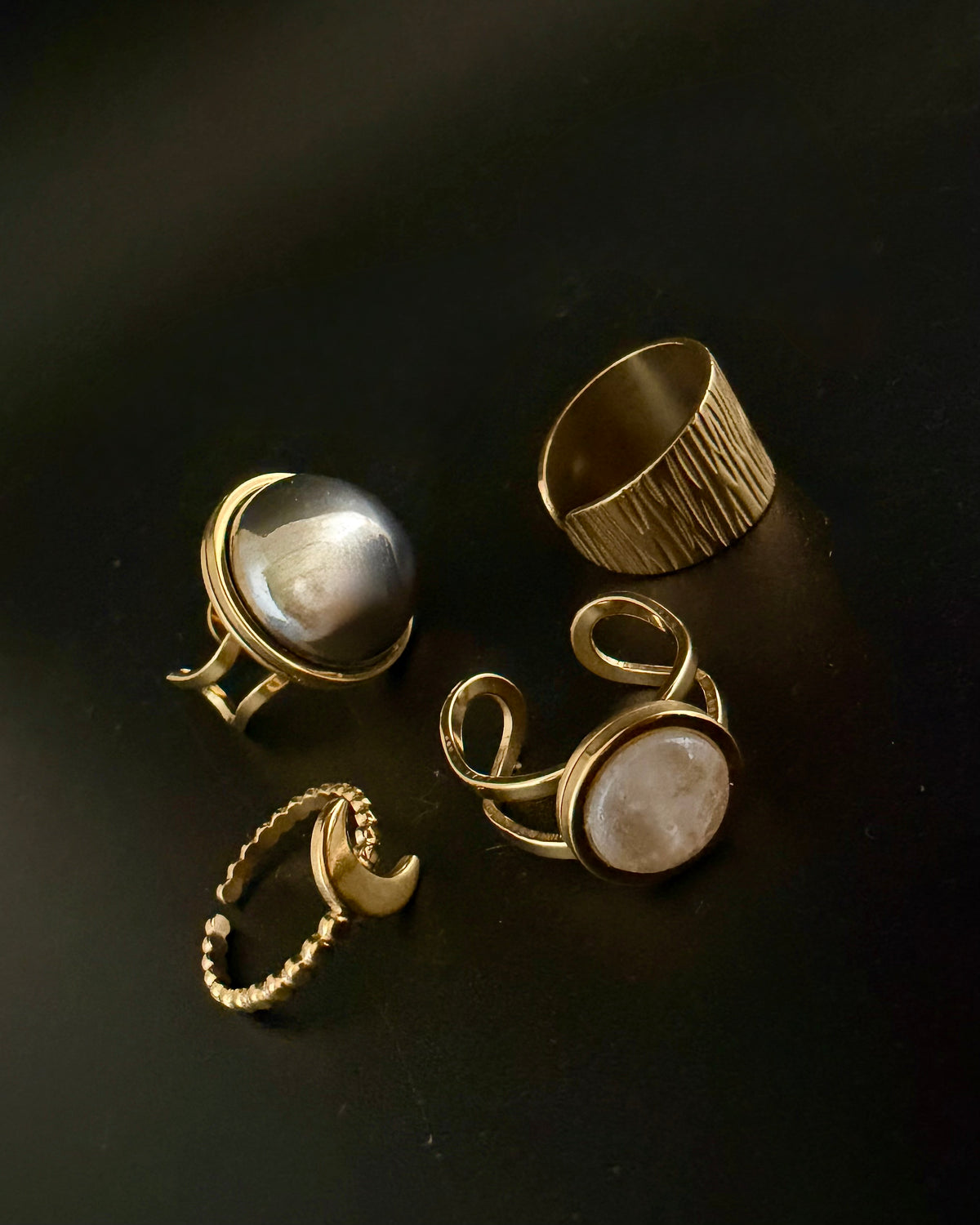 Four gold-toned rings displayed on a dark black background, including a pearl ring, a textured band ring, a beaded ring, and a ring with a pale stone.