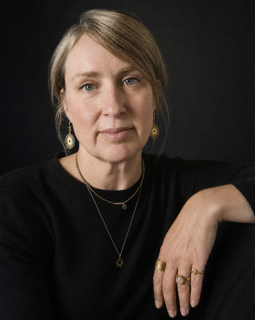 A close-up portrait of a woman with blonde hair tied back, wearing layered gold necklaces, matching sunburst earrings, and several gold rings, posed against a dark black background.