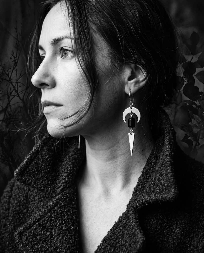 A black and white portrait of a woman in profile wearing a long, geometric dangle earring with a crescent moon, dark bead, and triangular drop, paired with a textured dark coat.