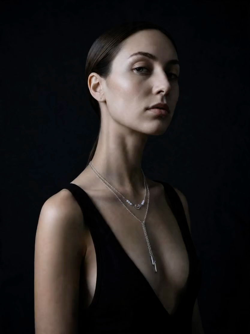 A woman in a black sleeveless top wearing a delicate silver Y-necklace with a long drop pendant, photographed against a dark black background.