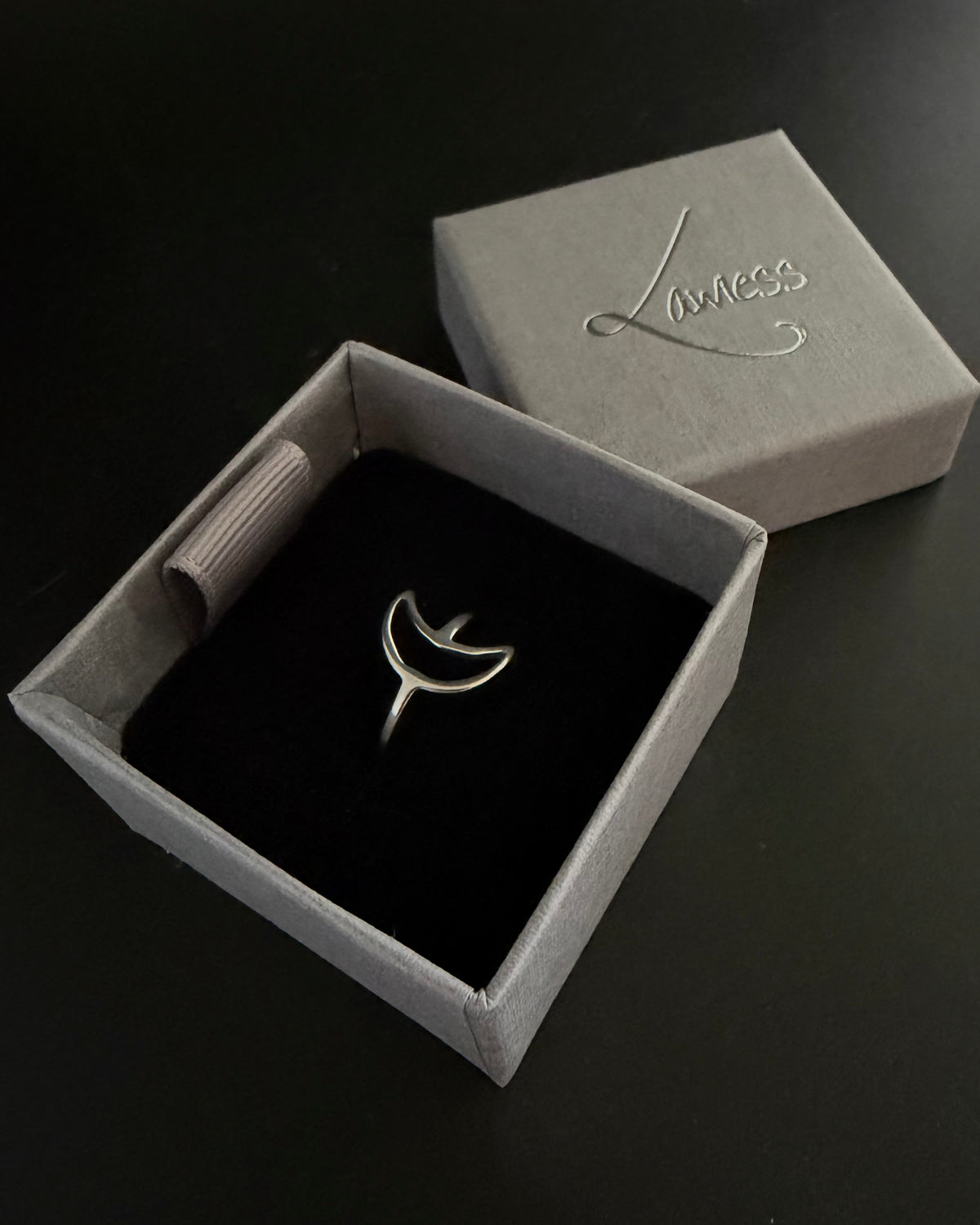 A silver crescent moon ring displayed inside an open gray jewelry box with a black velvet insert, with the matching gray lid placed behind it on a dark background.