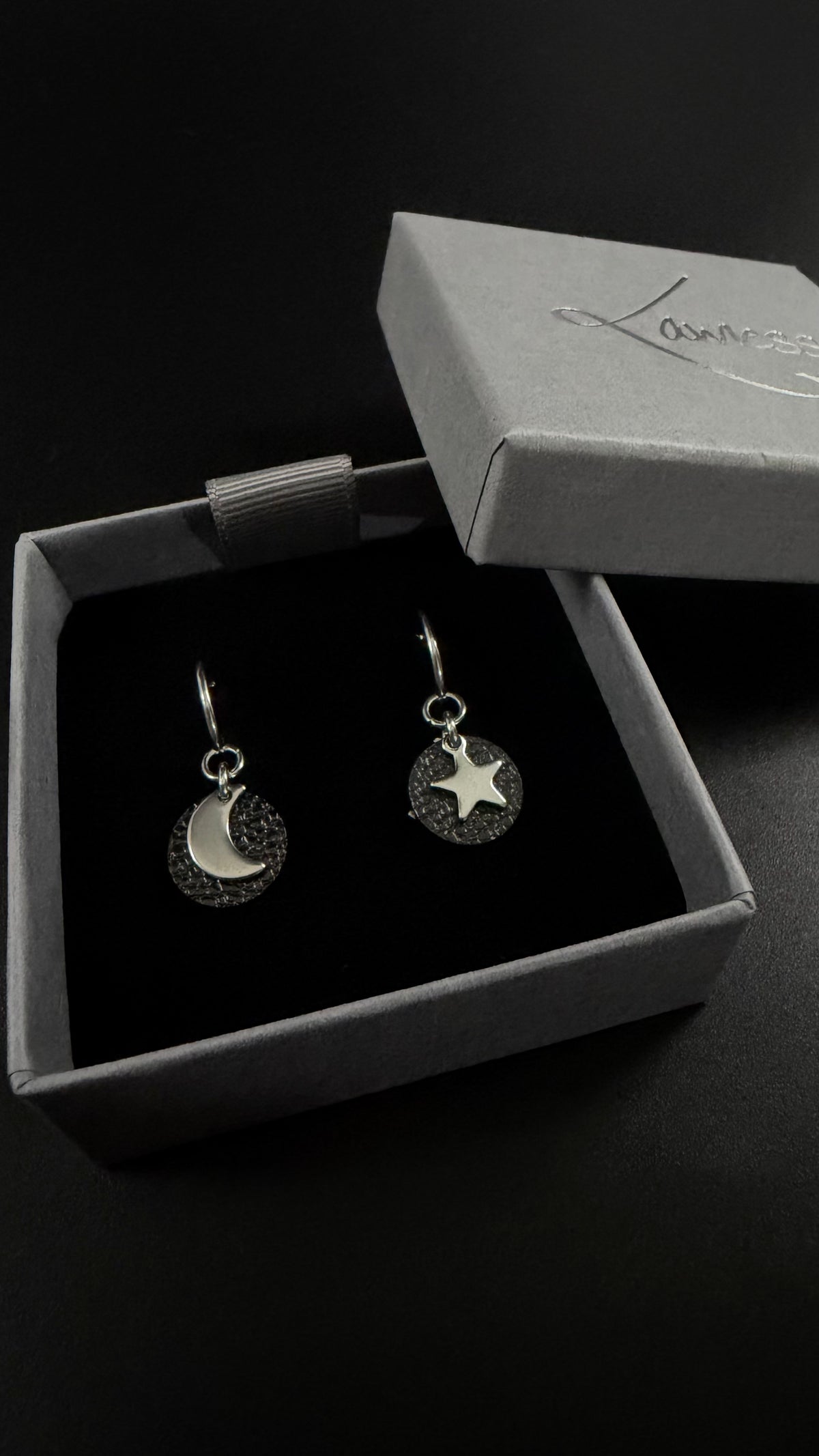 Urania Cipher silver hoop earrings with textured black reversible discs, shown with one silver crescent moon and one silver star against a dark background.
