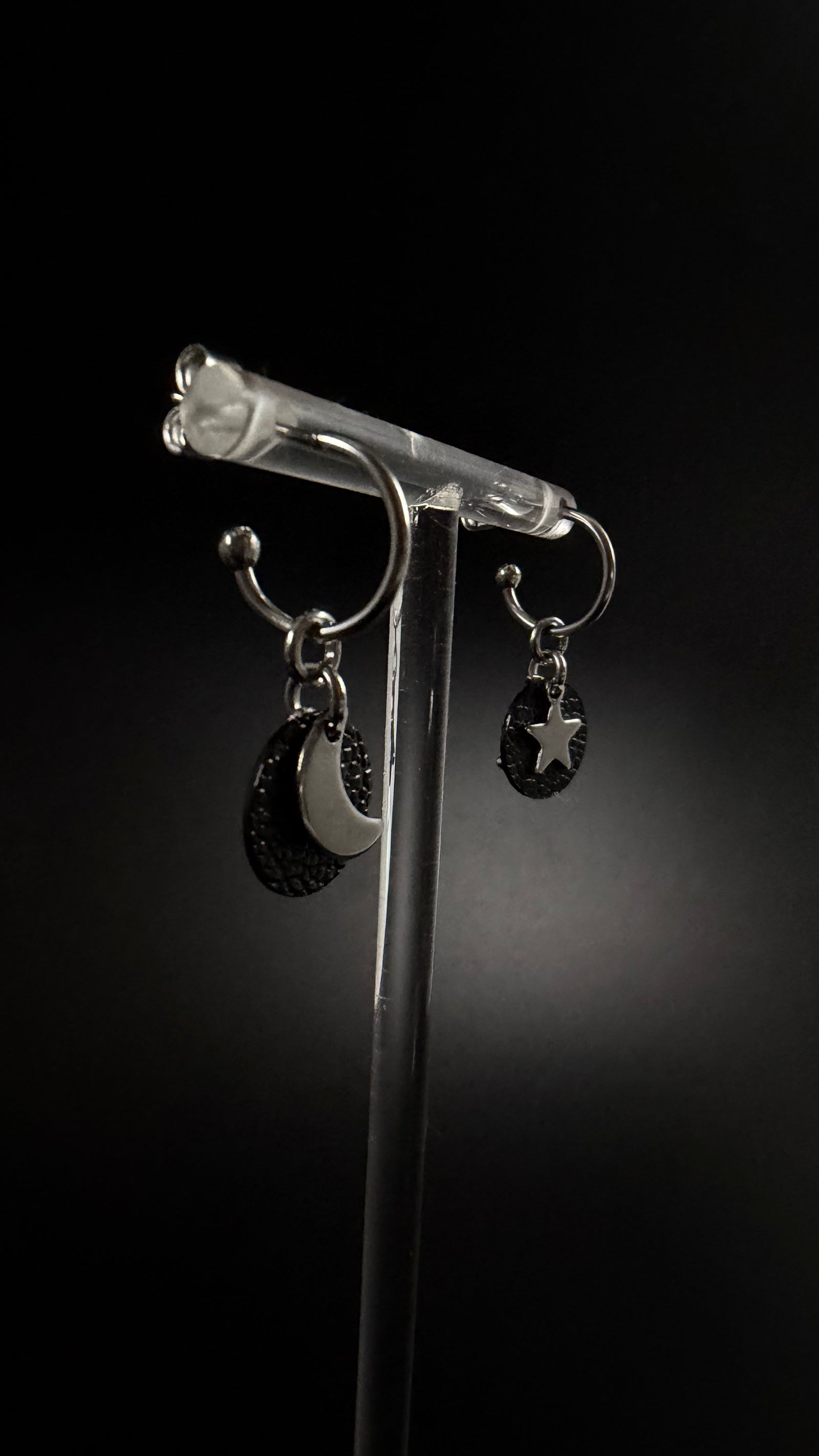 Urania Cipher silver hoop earrings with textured black reversible discs, shown with one silver crescent moon and one silver star against a dark background.